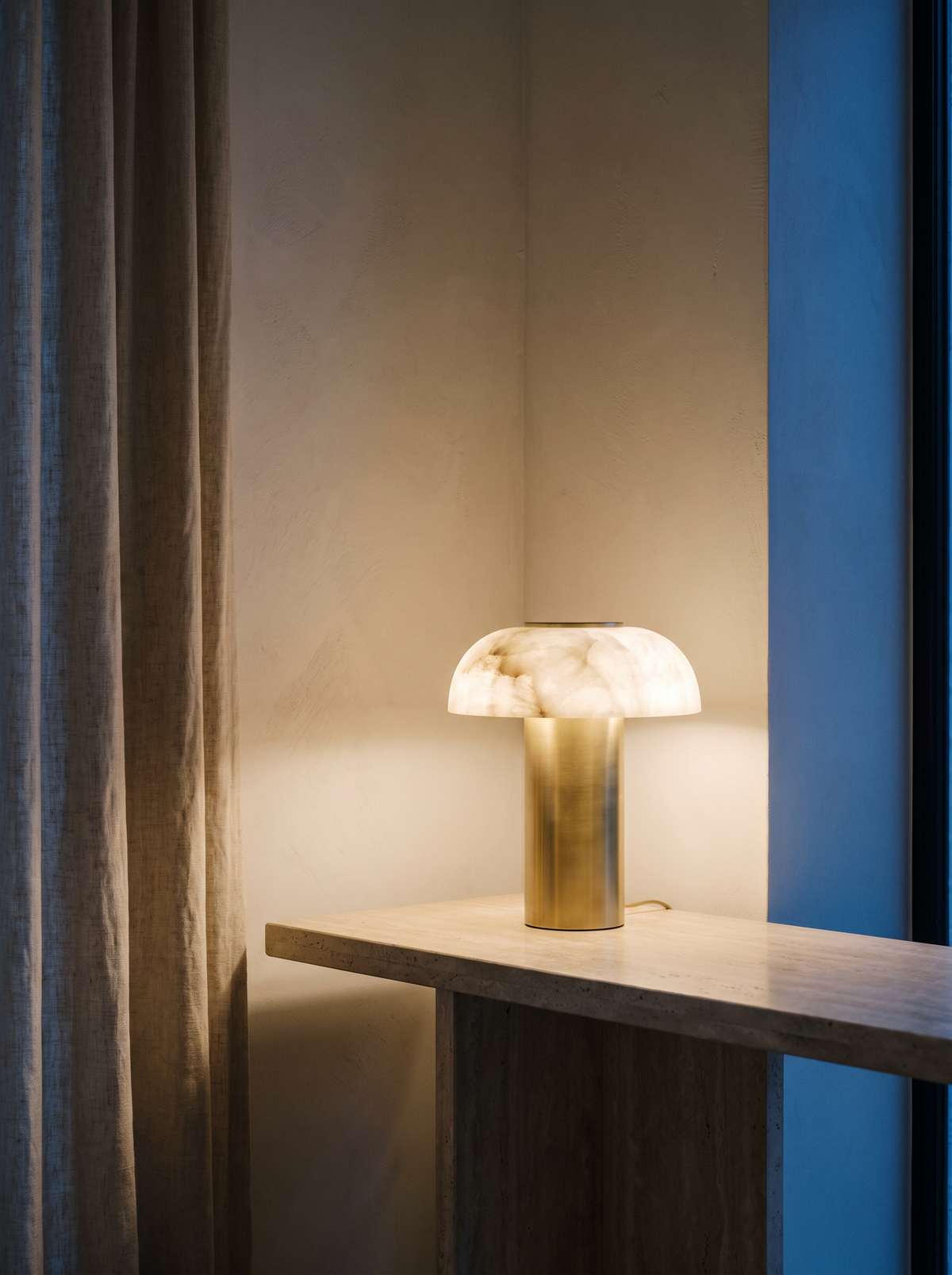 An alabaster and brass table lamp glowing on a travertine console at dusk