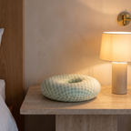 Small round donut ear pillow on a travertine bedside table beside a softly lit lamp