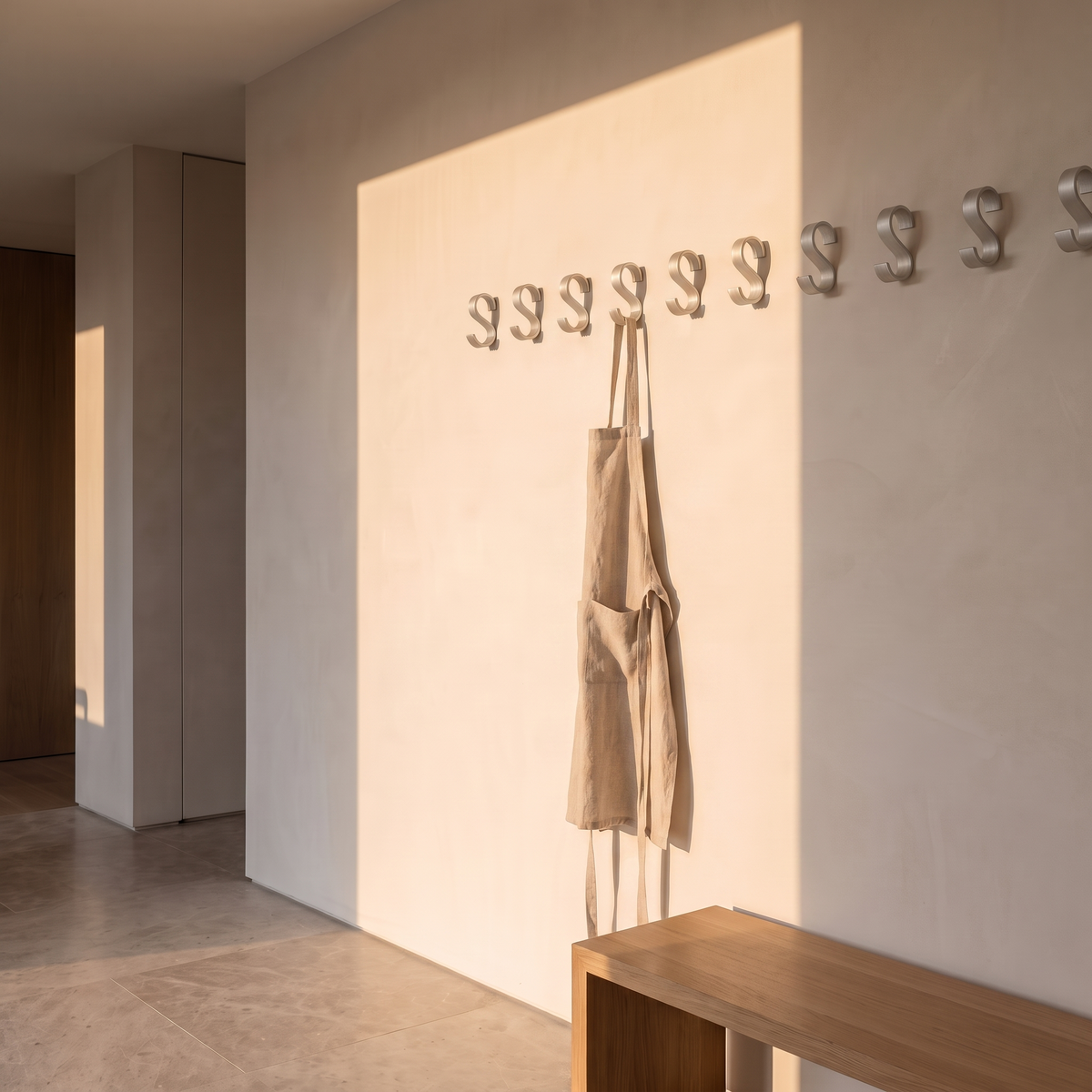 Set of matte aluminium wall hooks mounted on a plaster utility wall with a linen apron hanging from one